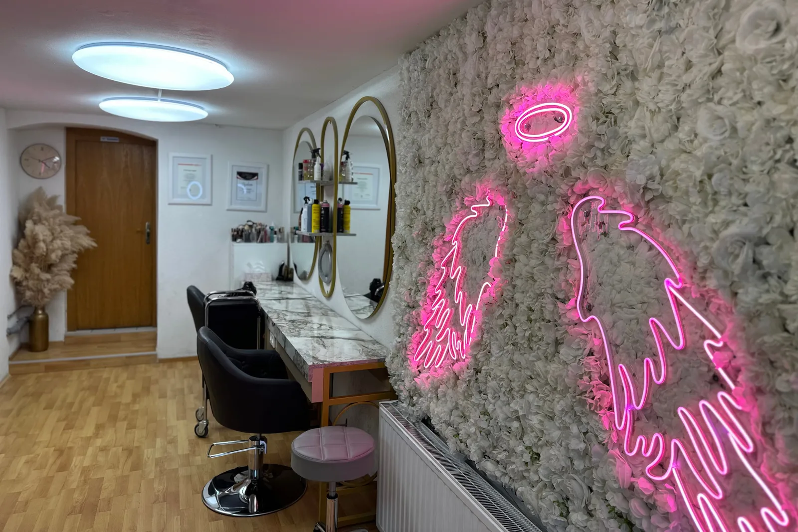 Salonraum von Hairlich: weiße Blütenwand mit pinkem Neon-Engelsflügel, goldgerahmte Ovalspiegel und Marmorkonsole